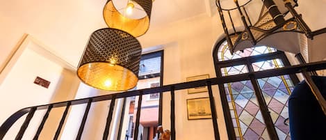 Un escalier avec une rampe noire, une statue en bois et une fenêtre ornée d’un motif en losange.