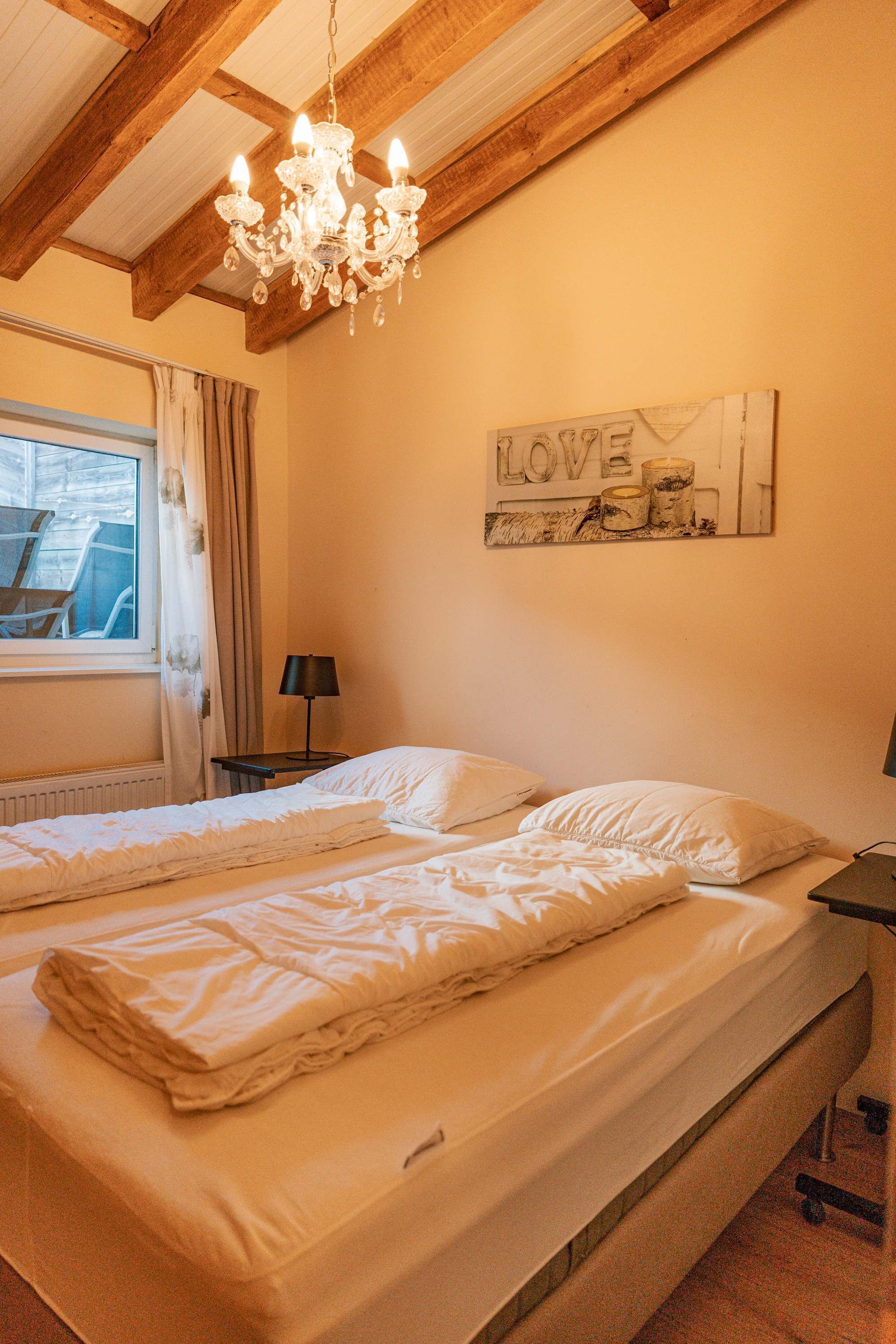 A double bed with white bedding, a chandelier, a window with curtains, and a wall decoration with the word 'LOVE'.