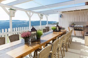Un área de comedor al aire libre techada, con una mesa de madera, sillas de mimbre, plantas en macetas y una parrilla.