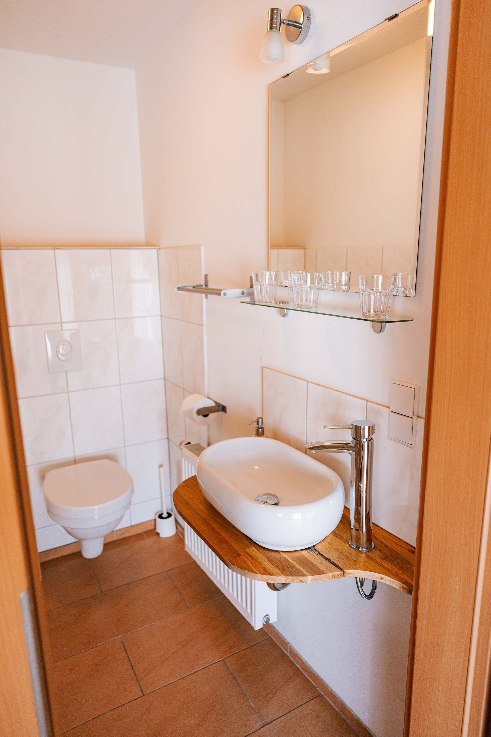 A bathroom with a white toilet, a white sink, a mirror, and a shelf with glasses.