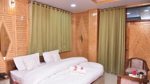 A hotel room with a large bed, a wooden headboard, green curtains, a wooden wall panel, and a small table with a chair. - Sarahan Holliday Inn (Rampur)