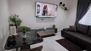En moderne stue med et fladskærms-tv på væggen, et spiseområde med bord og stole, og en mørk sofa.