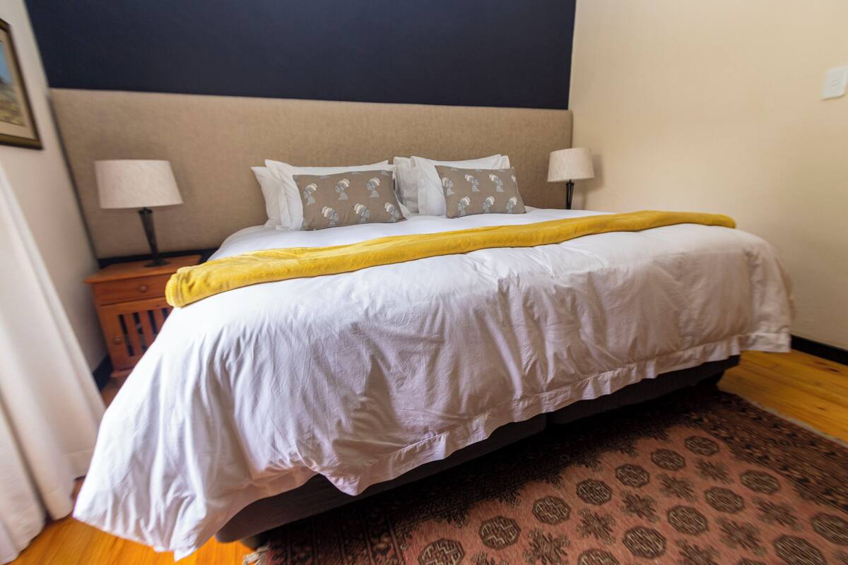 A hotel room with a bed, two bedside lamps, a patterned rug, and a dark wall.