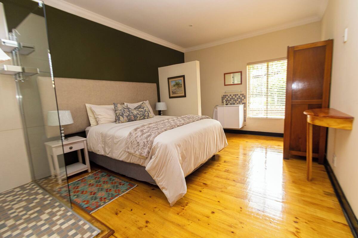 A bedroom with a bed, bedside tables, a nightstand, a wooden floor, a window with blinds, and a door.
