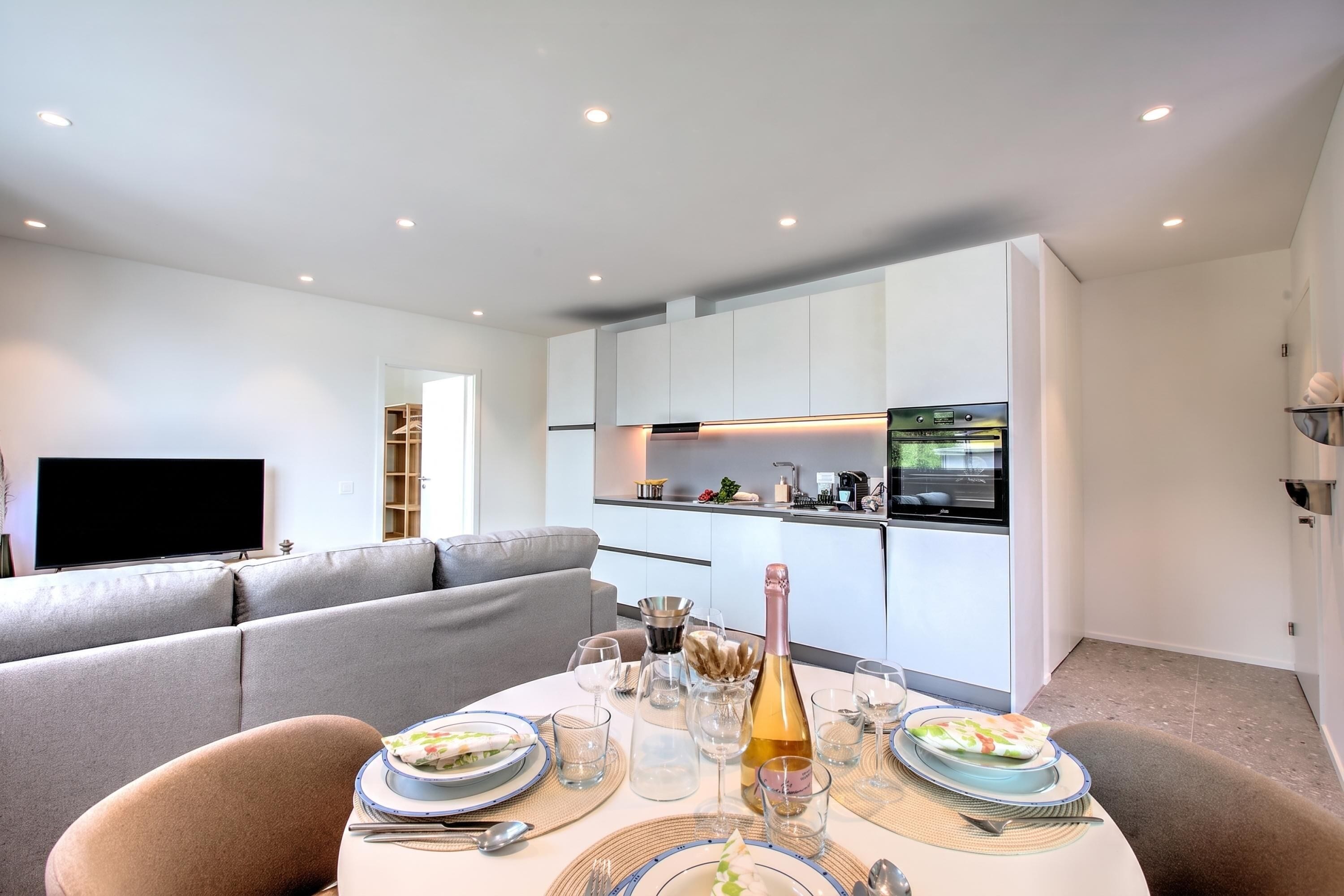 A modern living room with a dining area, a flat-screen TV, and a kitchen with white cabinets and a built-in oven.