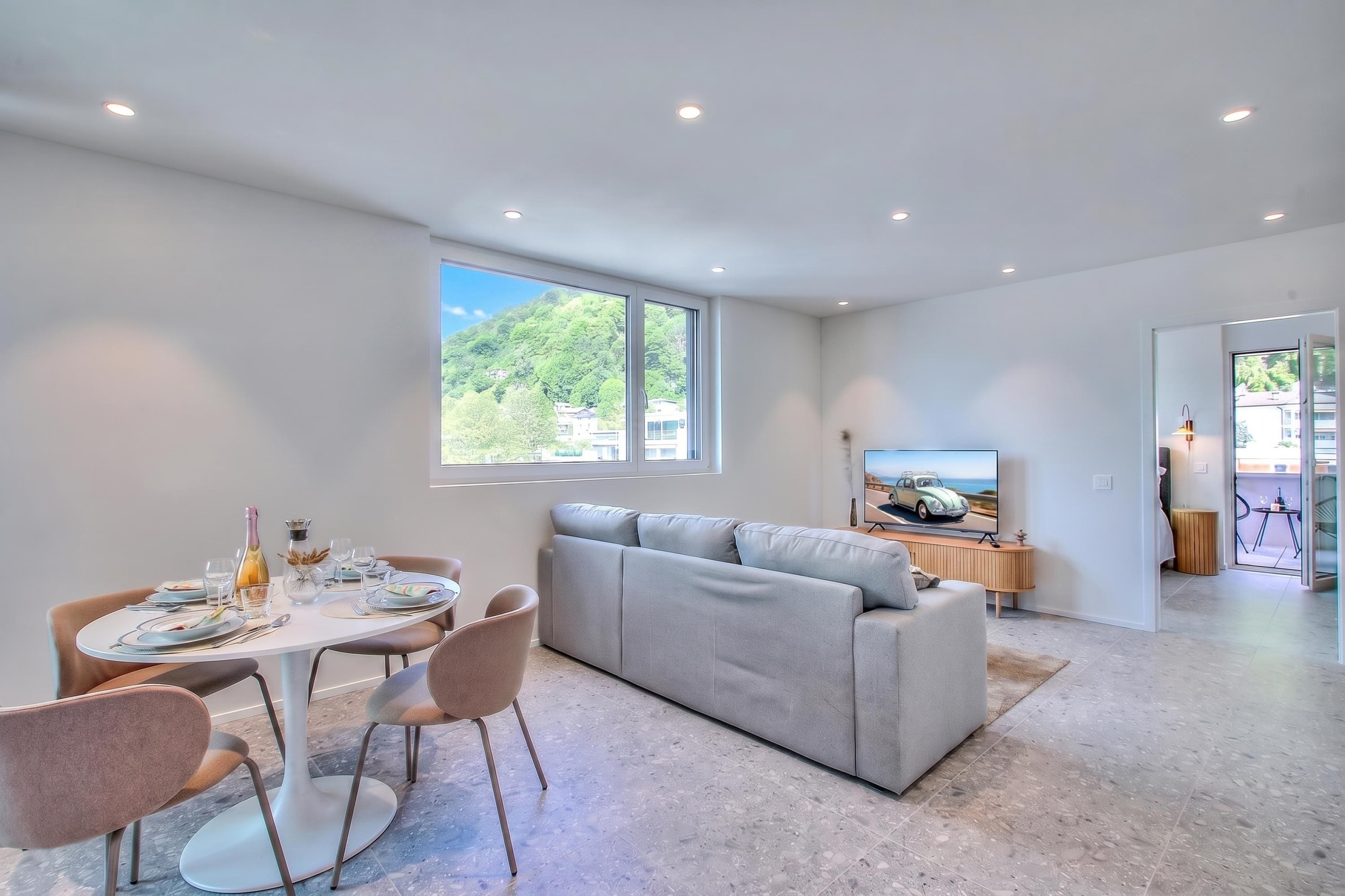 A modern living room with a grey sectional sofa, a dining table with chairs, and a television on a wooden stand.