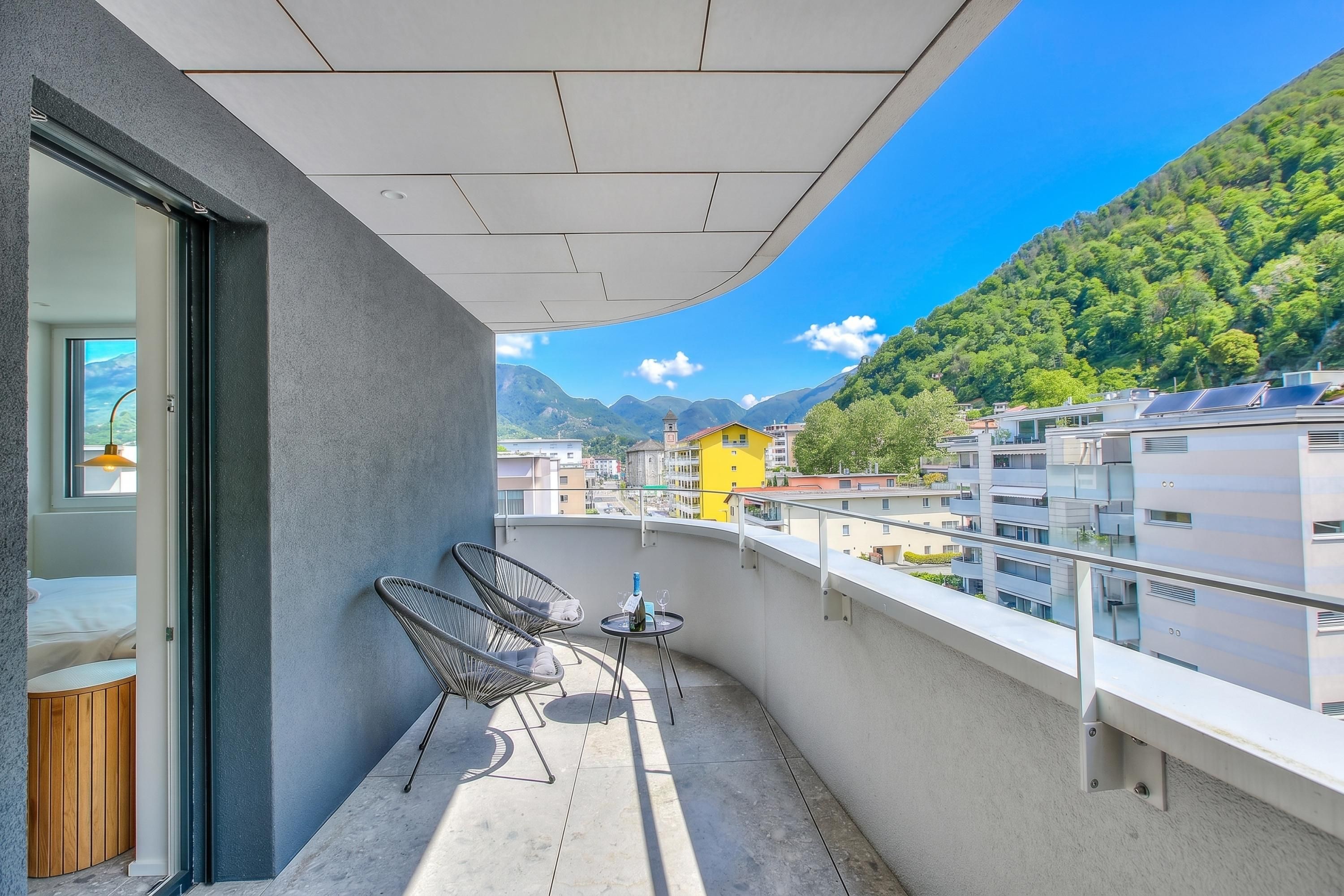 A balcony with a view of mountains, a yellow building, and a modern building with solar panels.