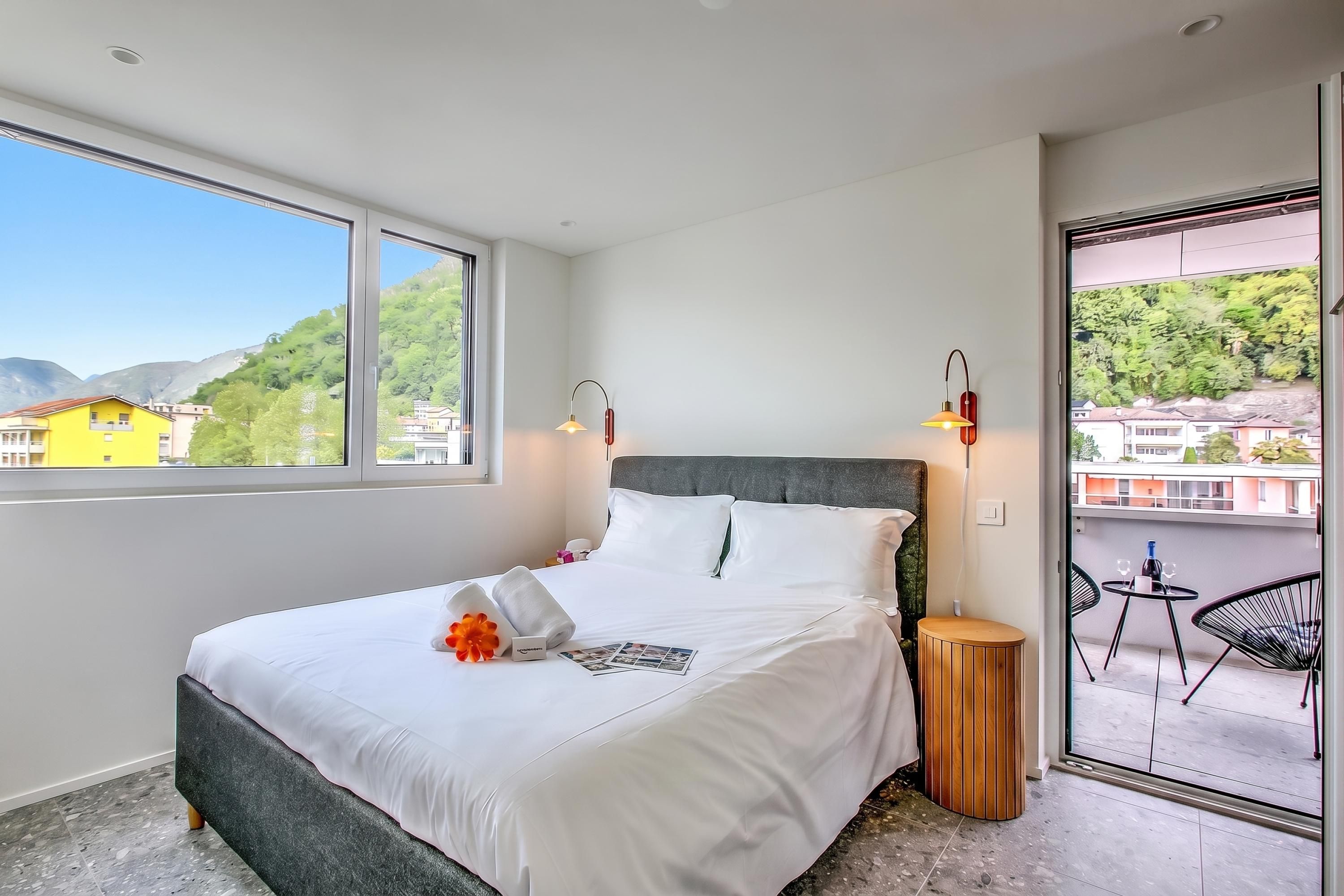 A hotel room with a large bed, two bedside lamps, a small wooden side table, and a balcony with a chair and table.