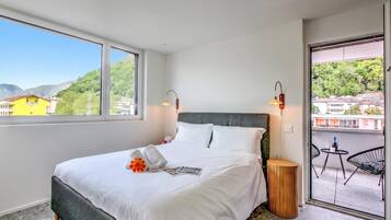A hotel room with a large bed, two bedside lamps, a small wooden side table, and a balcony with a chair and table.