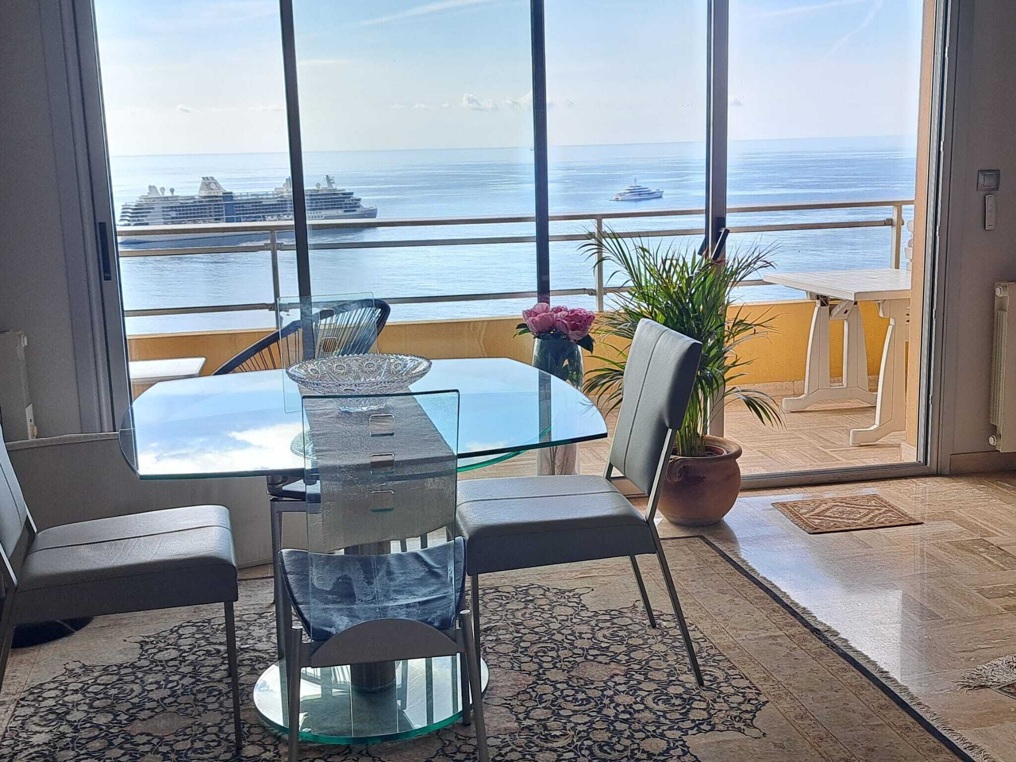 Een kamer met een glazen tafel en stoelen, een kamerplant en uitzicht op de oceaan en een cruiseschip.
