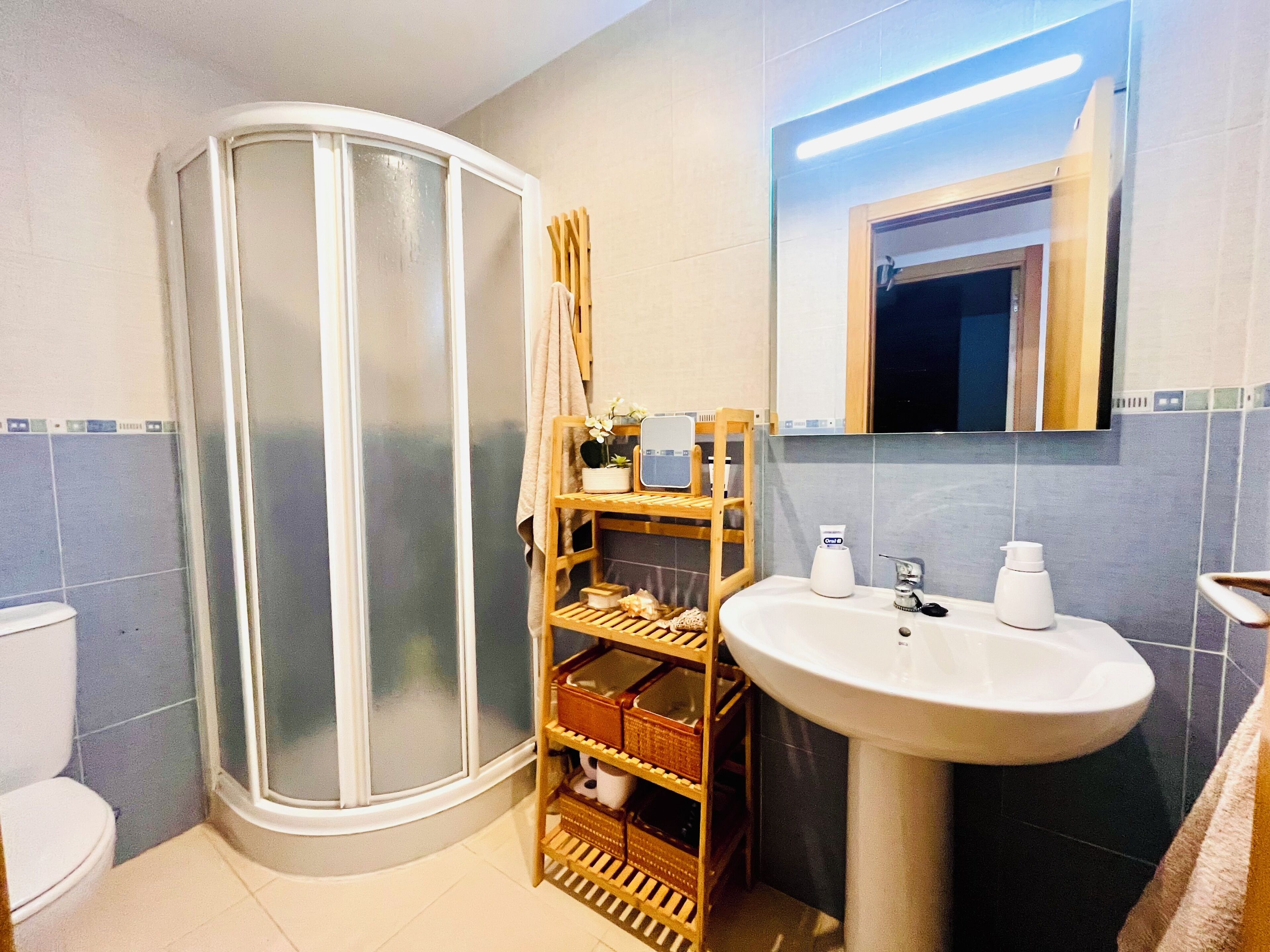 Une salle de bain avec une douche à parois de verre, une étagère en bois où sont rangés les articles de toilette, un lavabo blanc et un miroir.