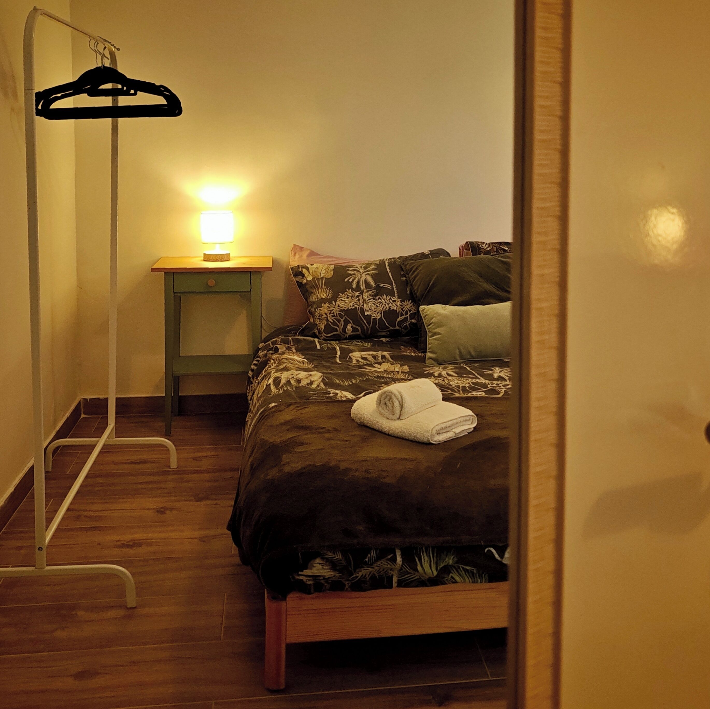 A bedroom with a bed, a bedside table with a lamp, a hanging clothes rack, and a wooden floor.