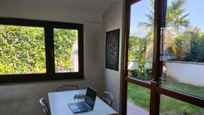 A room with a table, a laptop, and large windows with a view of greenery and a building.
