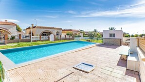 A swimming pool with a tiled deck, a small covered seating area, and a white building with a tiled roof.