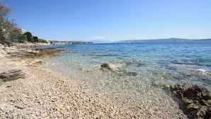 Una playa de guijarros con agua cristalina, una costa rocosa y montañas a lo lejos.
