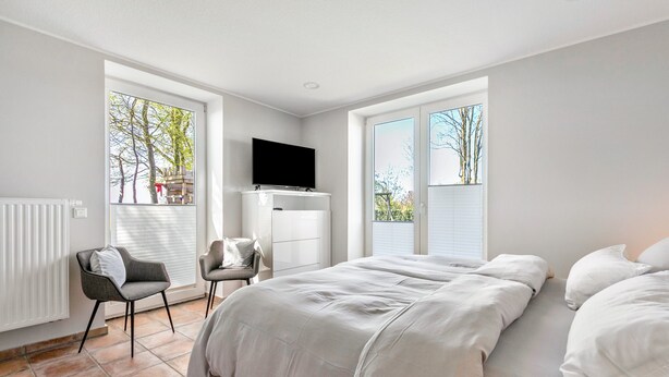 Ein modernes Schlafzimmer mit Bett, Sessel, Fernseher und großen Fenstern.