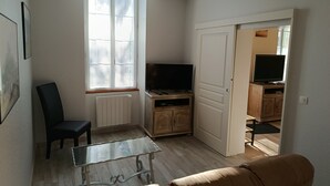 A living room with a glass coffee table, a sofa, a TV on a wooden cabinet, a radiator, and a window. - Between vineyards and estuary, discover Walter's winery (Semillac)