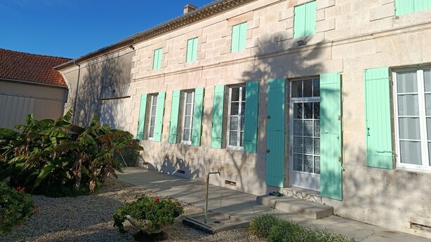 A two-story building with a stone facade and green shutters, featuring a small garden area with plants and a gravel path. - Between vineyards and estuary, discover Walter's winery (Semillac)