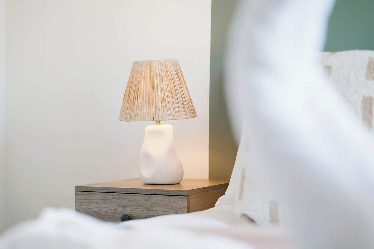 A bedside table with a lamp, a white vase, and a bed with white bedding.