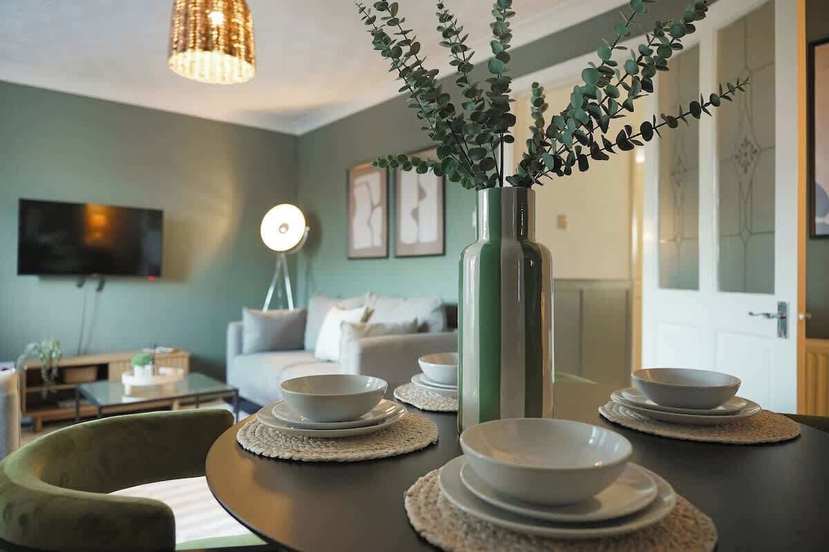 A dining area with a table set for four, a vase with greenery, a hanging light fixture, and a television in the background.