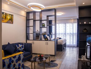 A modern living room with a sofa, a coffee table, a decorative screen, and a view of the outside through large windows. - Cozy,all-equipped 1-bedroom apartment with cafe,gym &minimart in Uptown Nairobi. (Nairobi)