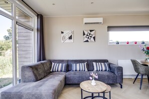 A modern living room with a grey sectional sofa, a round wooden coffee table, and a grey armchair. There are abstract wall art pieces and a white air conditioning unit.