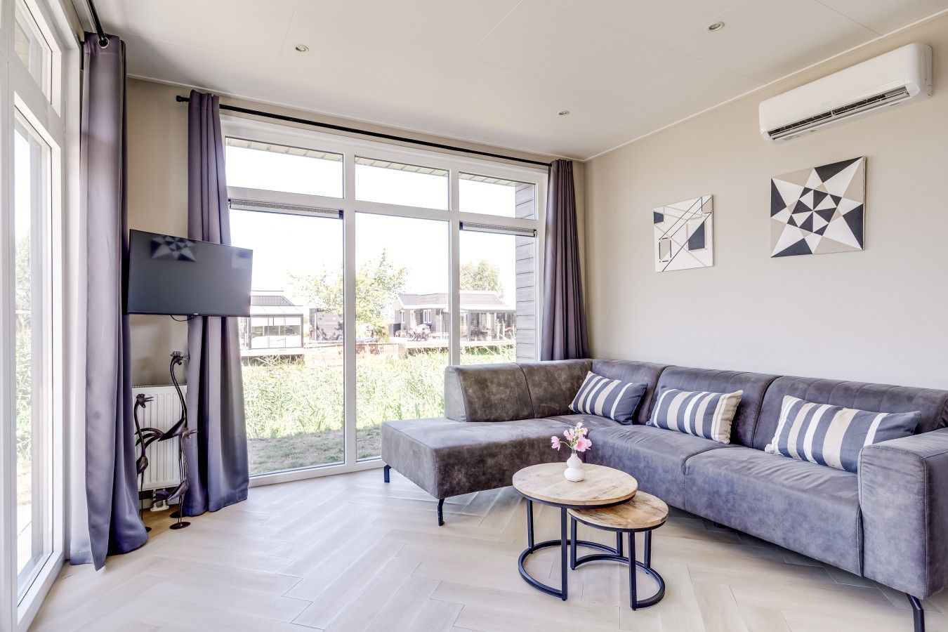 Een moderne woonkamer met een grijze bank, een ronde houten salontafel en een groot raam met gordijnen.
