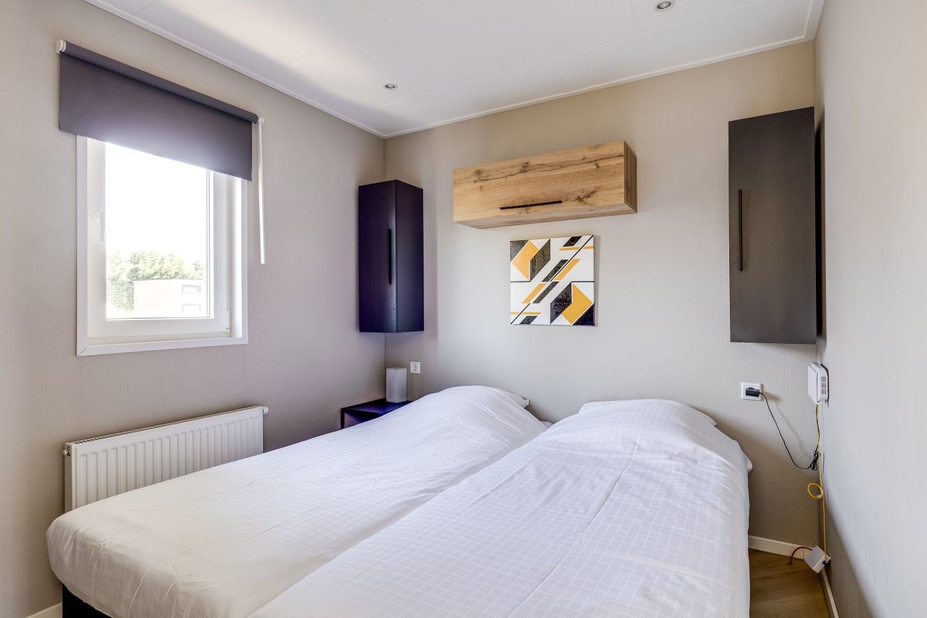 A bedroom with two beds, a window, a radiator, a wooden cabinet, a wall-mounted TV, and a power outlet.