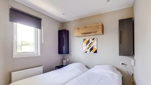 A bedroom with two beds, a radiator, a window with blinds, a wall-mounted TV, and a wooden shelf with a geometric artwork. - Cube XL 68 | 4 persoons chalet in Zennewijnen (Zennewijnen)