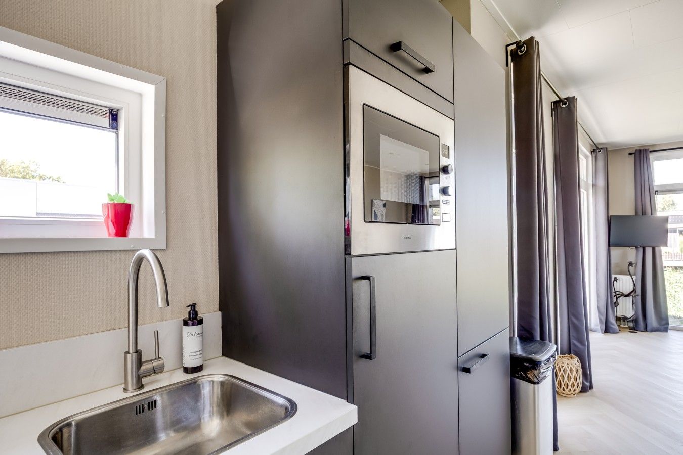A modern kitchen with a stainless steel sink, a built-in oven, and a window with a view of greenery.