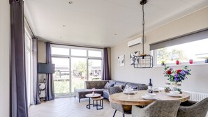 A modern dining area with a round table, grey upholstered chairs, and a view of the outdoors. - Cube XL 68 | 4 persoons chalet in Zennewijnen (Zennewijnen)