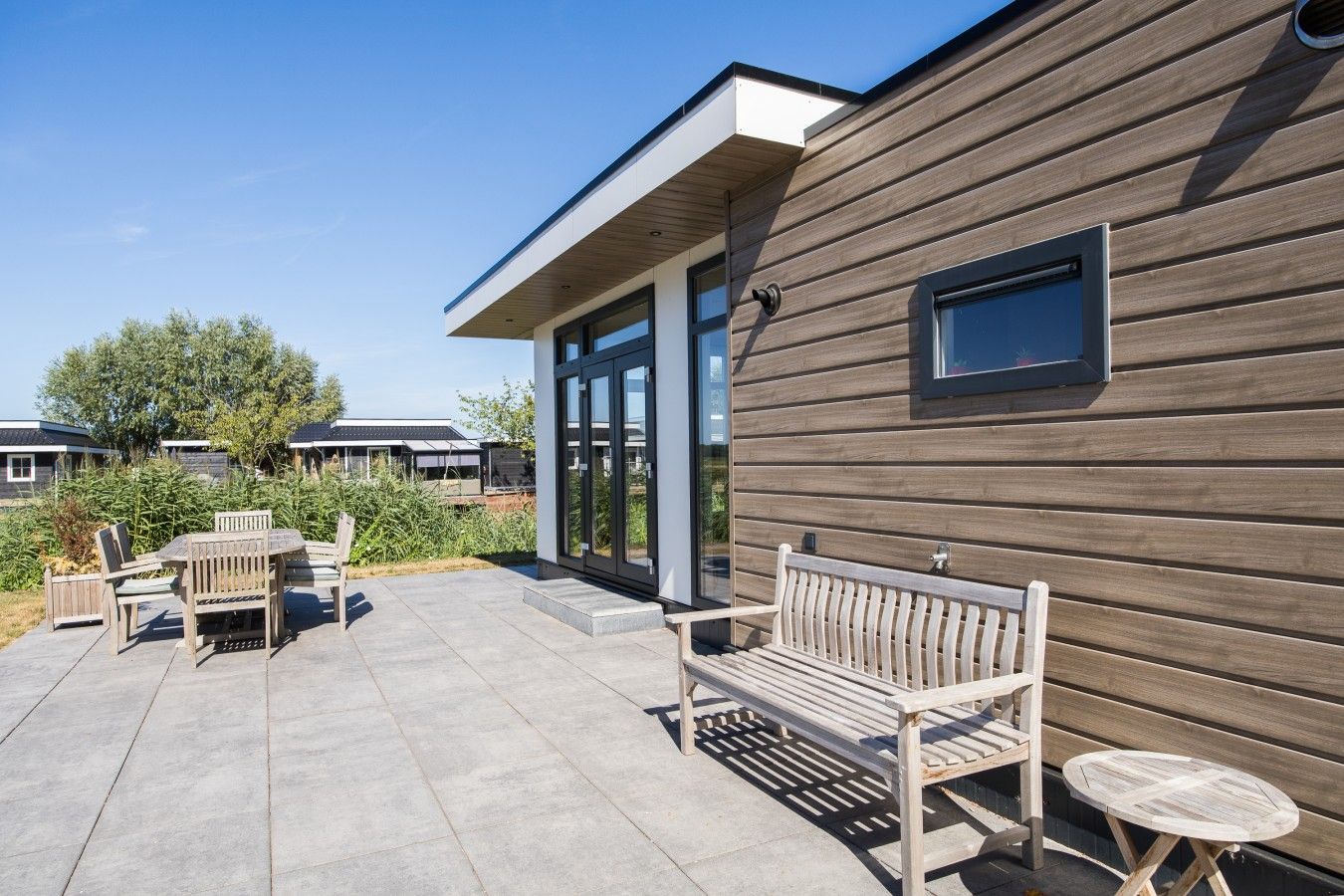 A modern building with a wooden exterior, a covered patio area with outdoor furniture, and a clear sky.