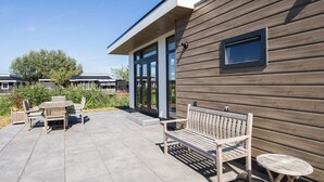 A modern building with a wooden exterior, a covered patio area with outdoor furniture, and a clear sky. - Cube XL 68 | 4 persoons chalet in Zennewijnen (Zennewijnen)