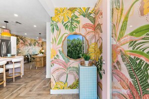 A room with a wooden floor, a dining area with a white table and chairs, a kitchen with a refrigerator, and a wall with a tropical leaf pattern. - Whimsical Palm Springs Retreat - A Ryson Property (Palm Springs)