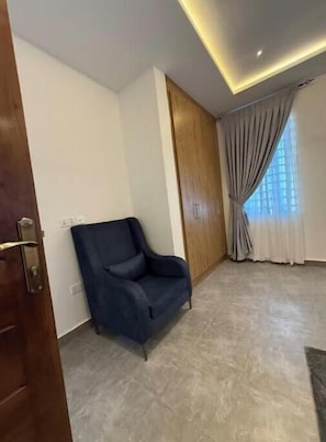 A room with a wooden door, a blue armchair, a wooden panel wall, a window with curtains, and a tiled floor.
