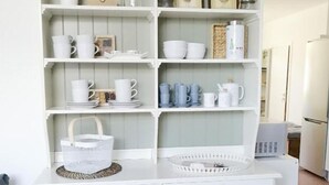 A white kitchen with open shelves containing various dishes and kitchenware, a small stool, and a refrigerator in the background.