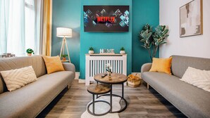 A living room with a sofa, a coffee table, a TV displaying the Netflix logo, a lamp, and a plant.