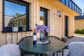 A modern building exterior with wooden siding, a window with plants, a table with a bottle and glass, and a sign. - Tiny Home “Auerberg Sweet” with Private Terrace and Wi-Fi (Stötten am Auerberg)