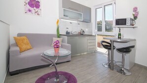 Een ruimtelijk ingerichte leefruimte met een kitchenette, een bank met een kussen, een kleine ronde tafel met een vaas en een paars vloerkleed.