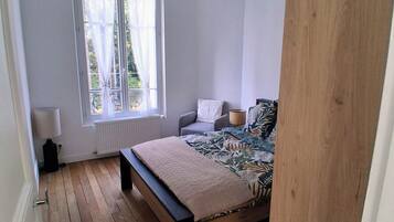 A bedroom with a wooden floor, a bed with a patterned bedspread, a window with curtains, and a ceiling light fixture.