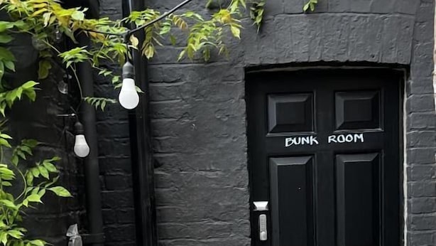 Una puerta negra con el rótulo 'BUNK ROOM' y una planta en maceta con flores rosadas en primer plano.