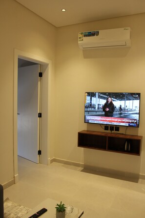Ein Wohnzimmer mit Flachbildfernseher an der Wand, einer kleinen Pflanze auf dem Boden und einer Fernbedienung.