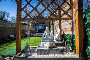 Un giardino con una pergola in legno, una statua di una figura seduta e una scultura a forma di testa di leone.