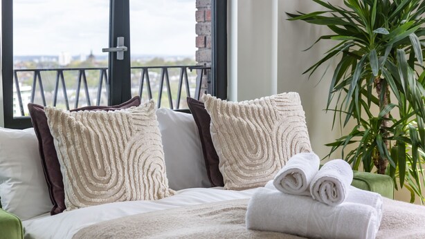 A bed with white and brown pillows, a rolled white towel, and a view of a balcony with a railing. - Modern 1BR Kings Cross Hideaway (London)