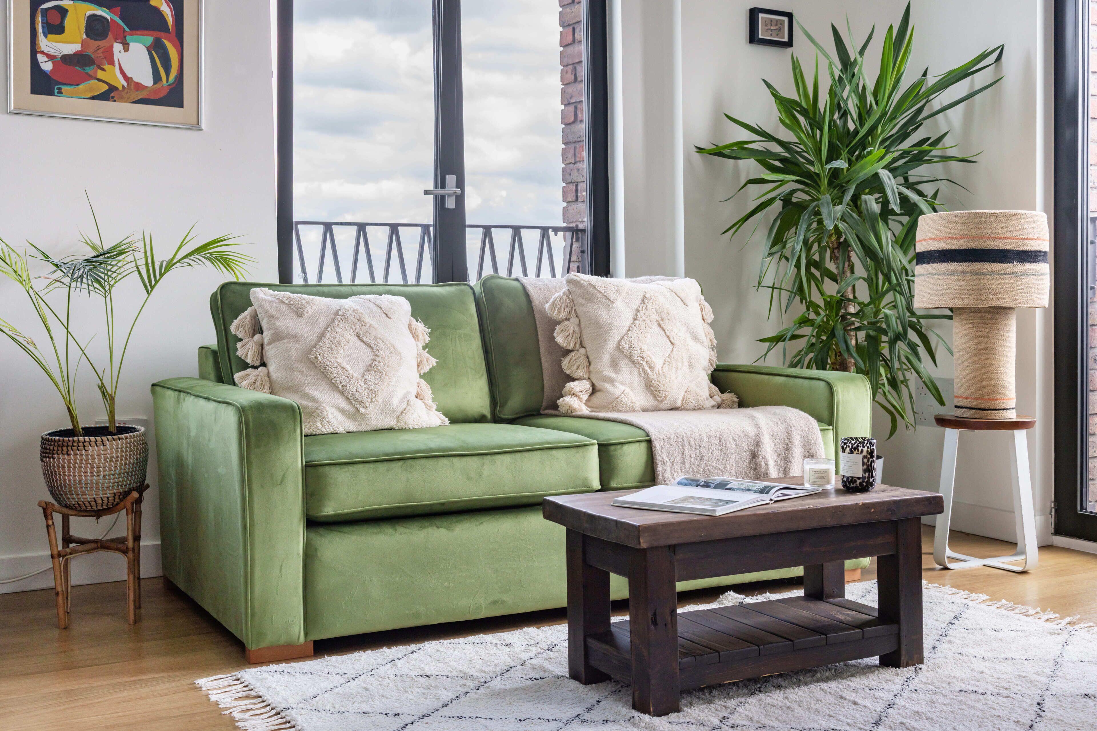 A green sofa with white throw pillows, a wooden coffee table, a wicker plant pot, and a tall potted plant.
