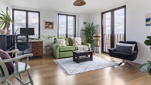 A spacious living room with a green sofa, a black armchair, a wooden coffee table, and a white chair. There are large windows, a balcony, and potted plants. - Modern 1BR Kings Cross Hideaway (London)