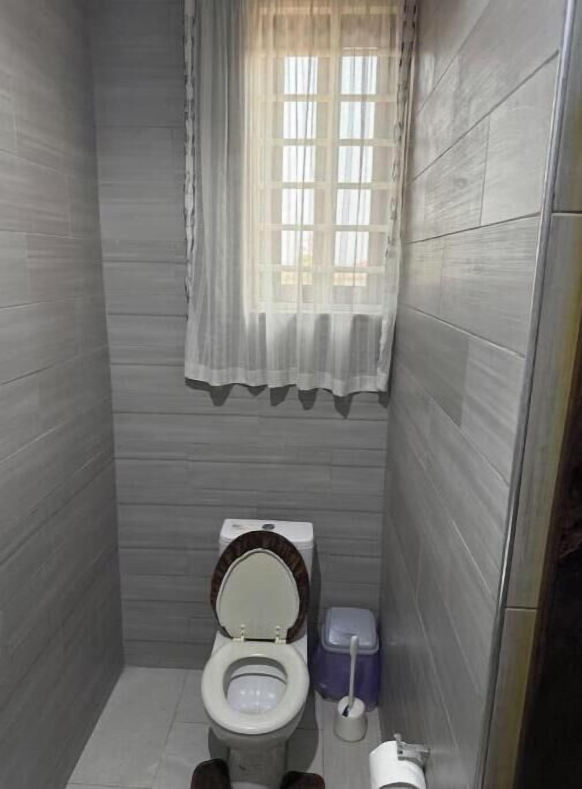 Une petite salle de bain étroite, avec des toilettes, une poubelle et une fenêtre ornée d’un rideau léger.