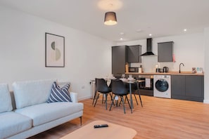 Ein modernes Wohnzimmer mit einer Couch, einem Esstisch mit Stühlen, einer Küchenzeile inklusive Schränken und einer Waschmaschine, sowie einer Pendelleuchte.