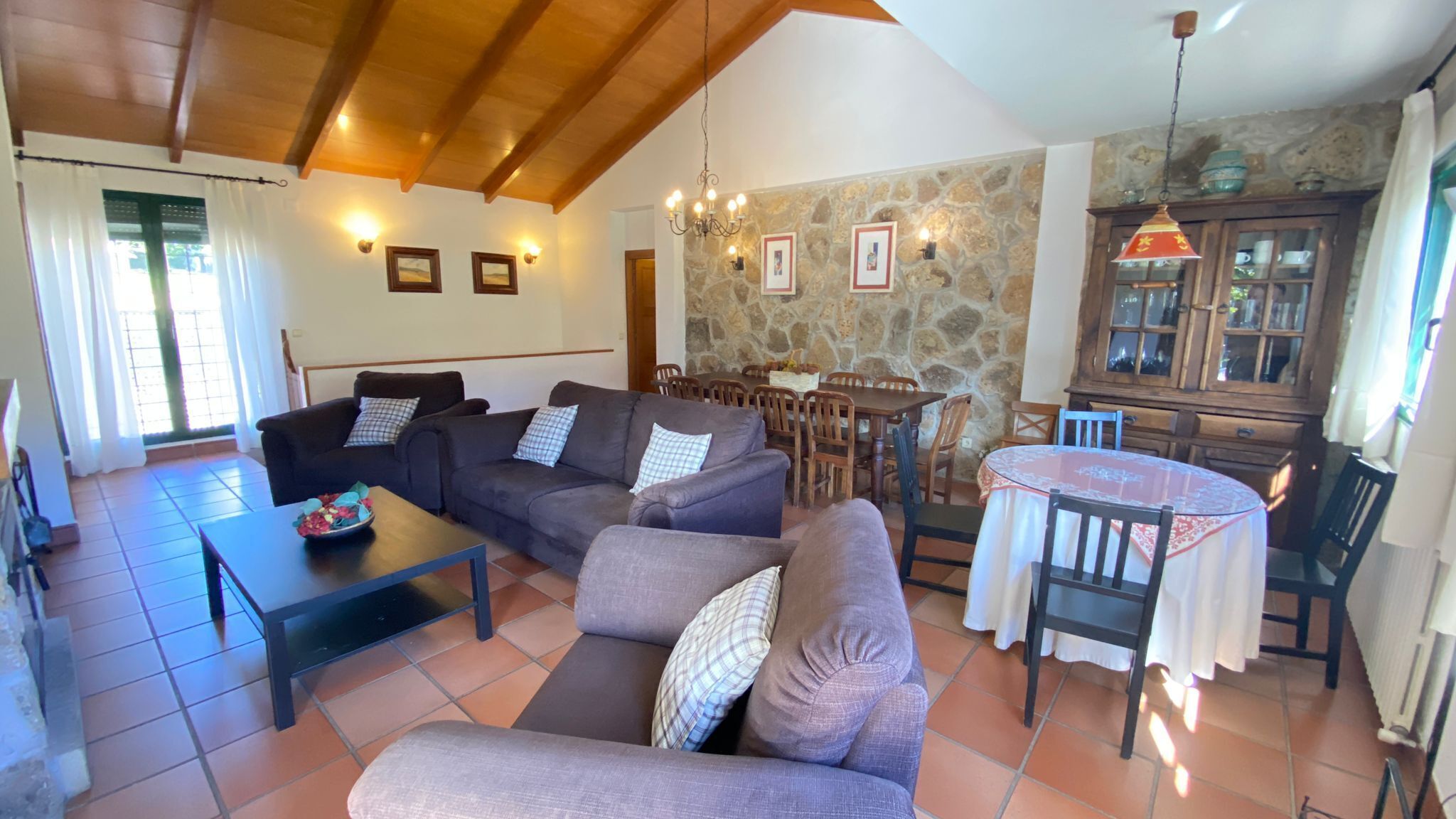 Una sala de estar con sofás, una mesa de centro, una zona de comedor con una mesa y sillas, una cocina con armarios y una pared de piedra.
