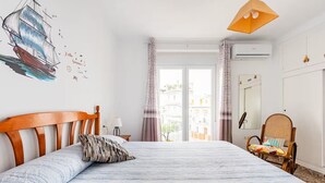 Ein Schlafzimmer mit einem Bett, einem Holz-Kopfteil, einem Rattan-Sessel, einem Fenster mit Vorhängen und Blick auf Gebäude.